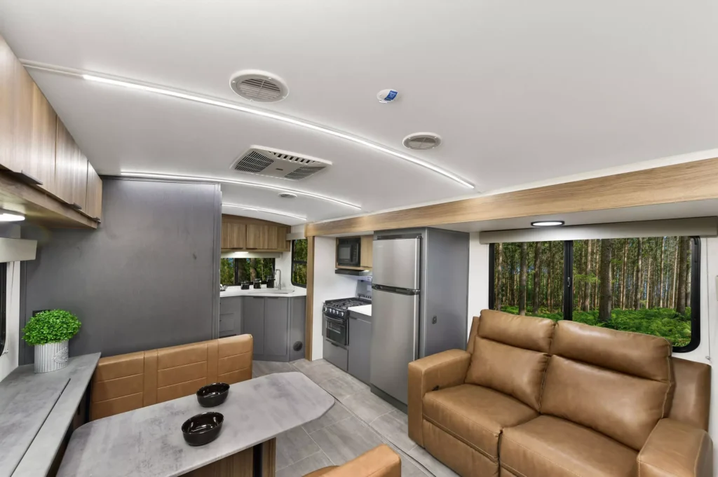 Modern RV interior with tan leather couch, matching dining benches, small dining table, kitchen area with stainless steel appliances, potted plant, and large windows showing a view of trees outside.
