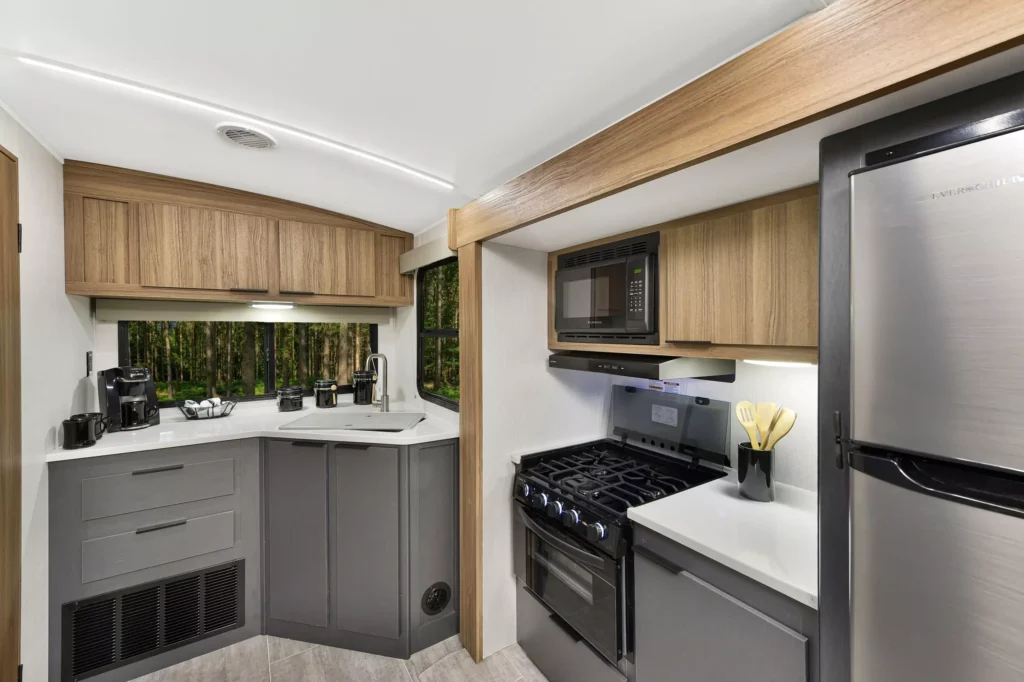 Modern RV kitchen with gray cabinets, stainless steel refrigerator, gas stove, microwave, and coffee maker. Large windows offer a view of green trees, and countertops display utensils and other kitchen essentials.