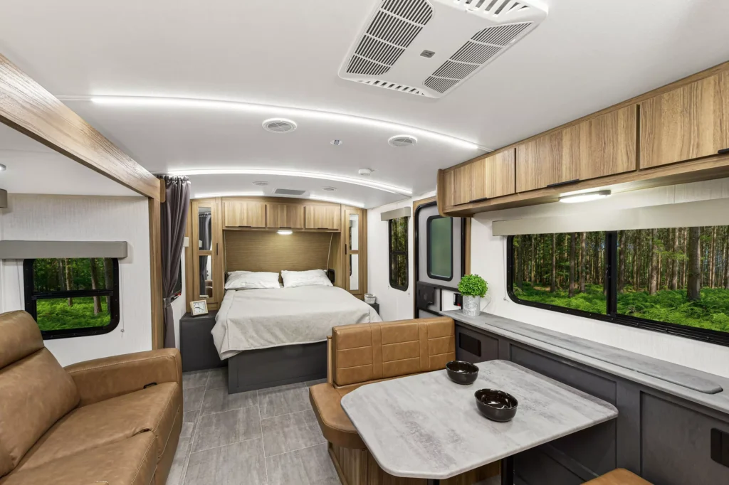 Modern RV interior with a bed, table, sofa, overhead cabinets, and large windows showing a forest view. The space is well-lit with LED lighting and features neutral tones and contemporary decor.