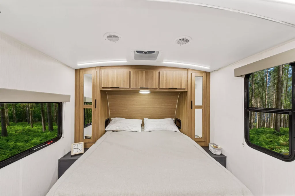A cozy RV bedroom with a neatly made bed, wood cabinets above, nightstands on each side, and large windows showing a green forest outside. The interior is bright and modern.
