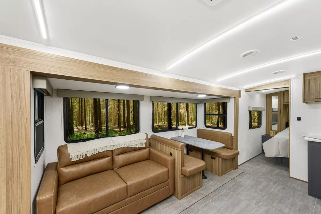 Modern RV interior with brown leather sofa, dining table with bench seating, wood accents, large windows showing a forest, and a view into the kitchen and bedroom areas.