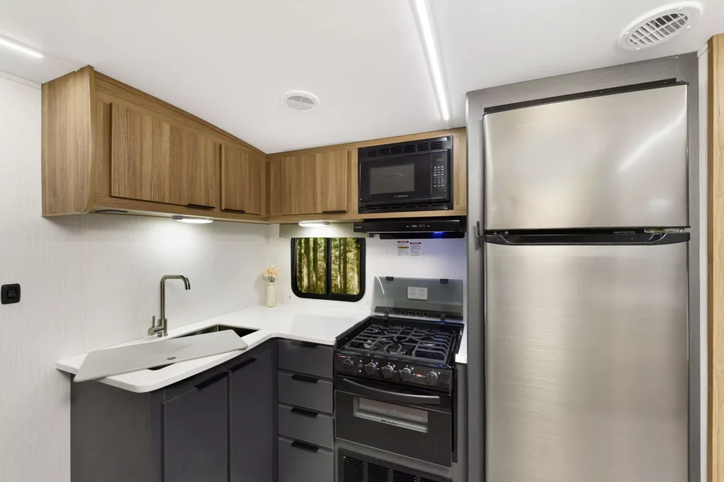 Modern RV kitchen with wood cabinets, stainless steel fridge, black stove with oven, mounted microwave, a white sink, and a window above the sink. The space is clean, compact, and well-lit.