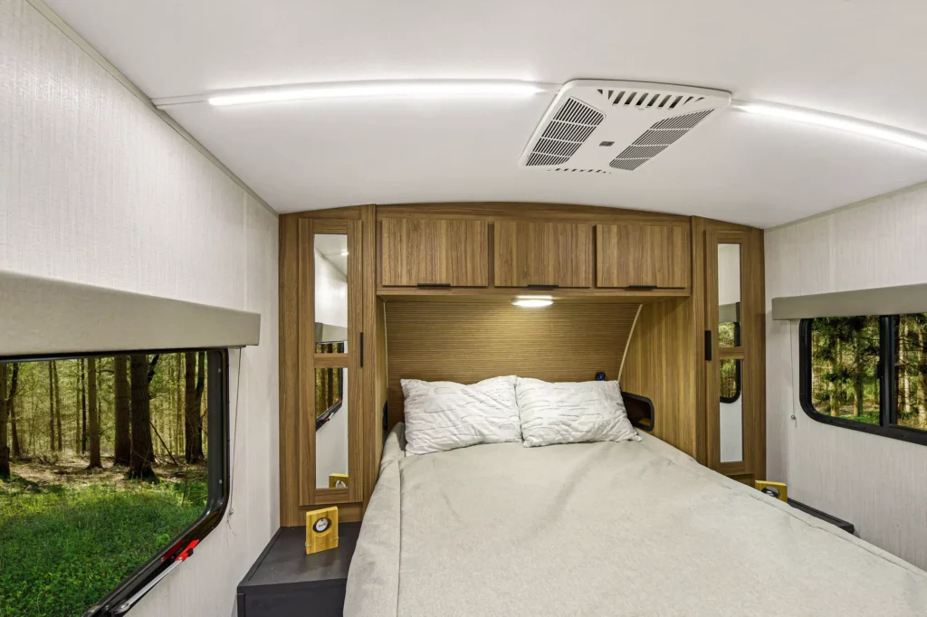 A cozy bedroom in a camper van with a double bed, white bedding, wooden storage cabinets above, mirrors on both sides, and windows showing a green forest outside. Ceiling lights and an air vent are visible above.