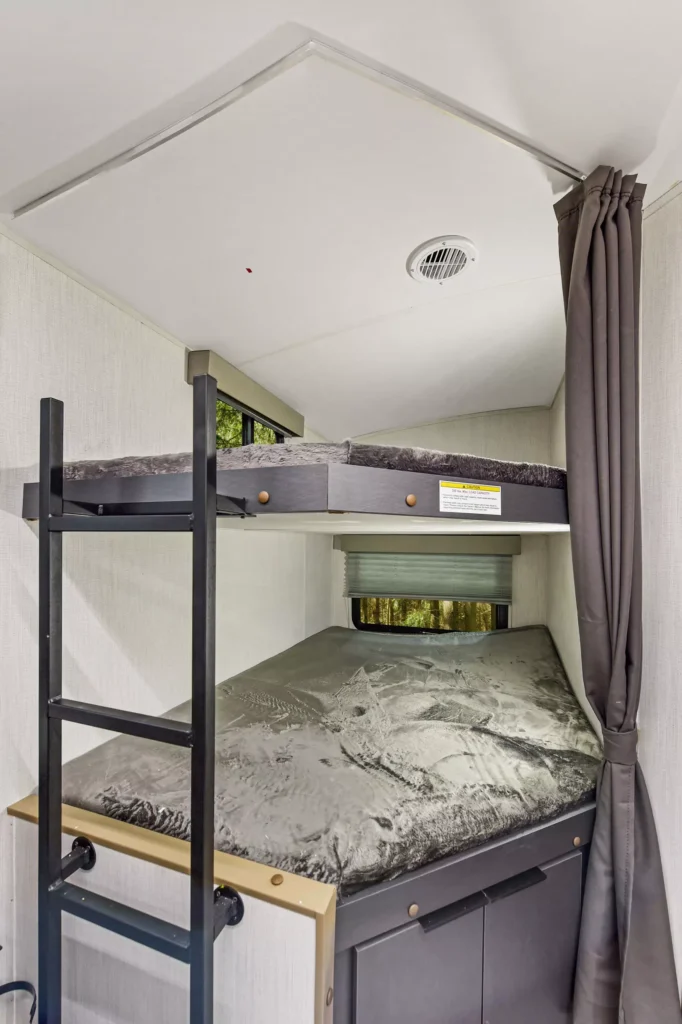 A compact bunk bed setup inside a camper with a ladder leading to the top bunk, a curtain for privacy, and storage cabinets beneath the lower bed.