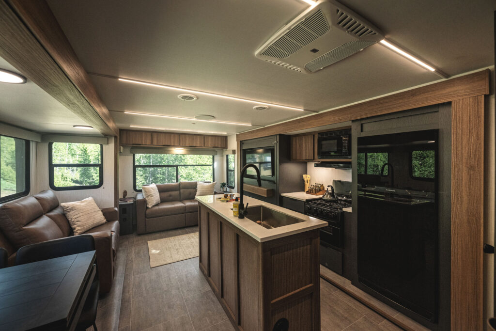 Modern RV interior featuring a kitchen island with a sink, stove, oven, microwave, large fridge, brown leather sofas, and large windows with views of greenery outside. The space is well-lit and stylishly furnished.