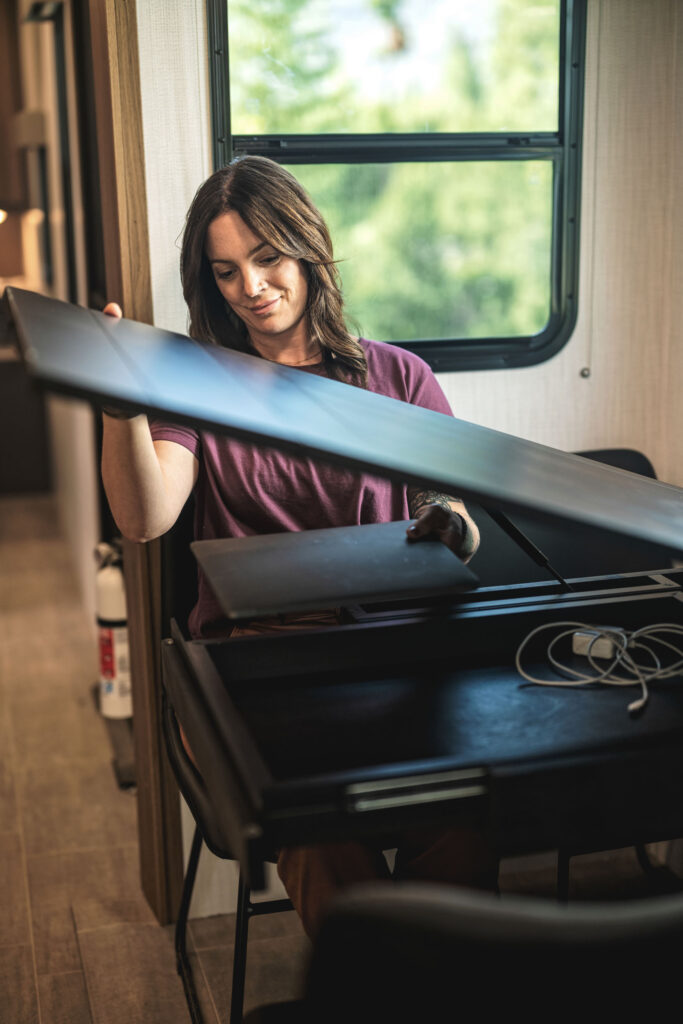 A woman sits at a desk by a window, lifting the lid of a black desk organizer. Earbuds and a charging cable are visible on the desk. Sunlight streams in through the window, and a fire extinguisher is in the background.