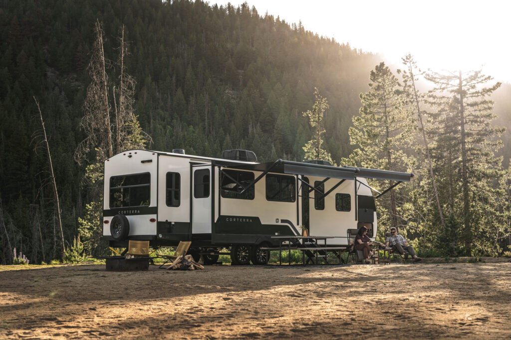 A large RV with an extended awning is parked in a scenic mountain clearing. Three people sit on chairs by a campfire, surrounded by tall pine trees and soft sunlight filtering through the forest.