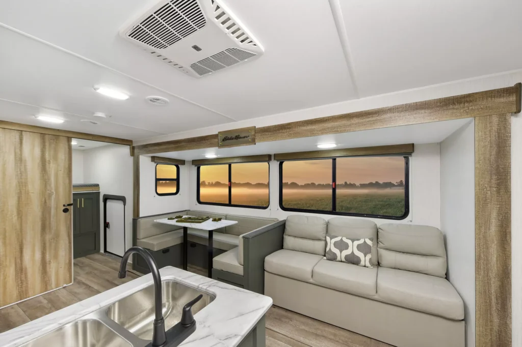Interior of an RV featuring a beige sofa with patterned pillows, a small dining booth, and a marble countertop with a sink. Large windows show a scenic sunset over a grassy field.