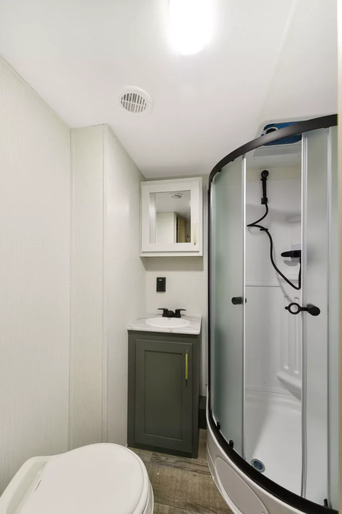 A compact bathroom features a toilet, a small sink with a cabinet and mirror above, and a curved glass shower enclosure with black fixtures. The walls are light-colored, and the space appears clean and modern.