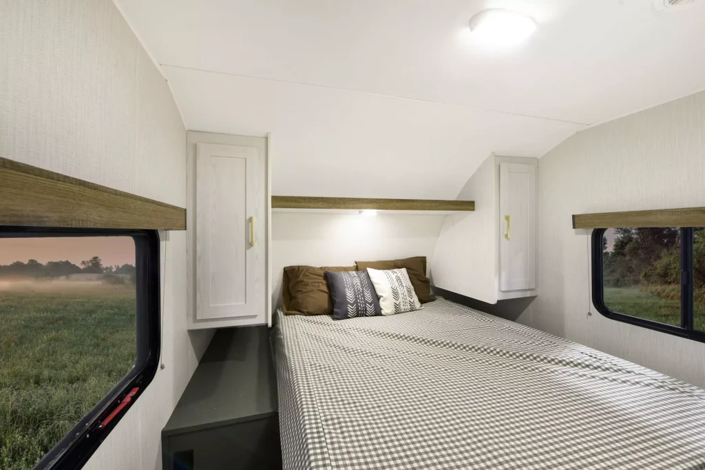 A cozy camper bedroom with a checkered bedspread, three decorative pillows, two overhead cabinets, and large windows showing a scenic outdoor view of grass and trees.