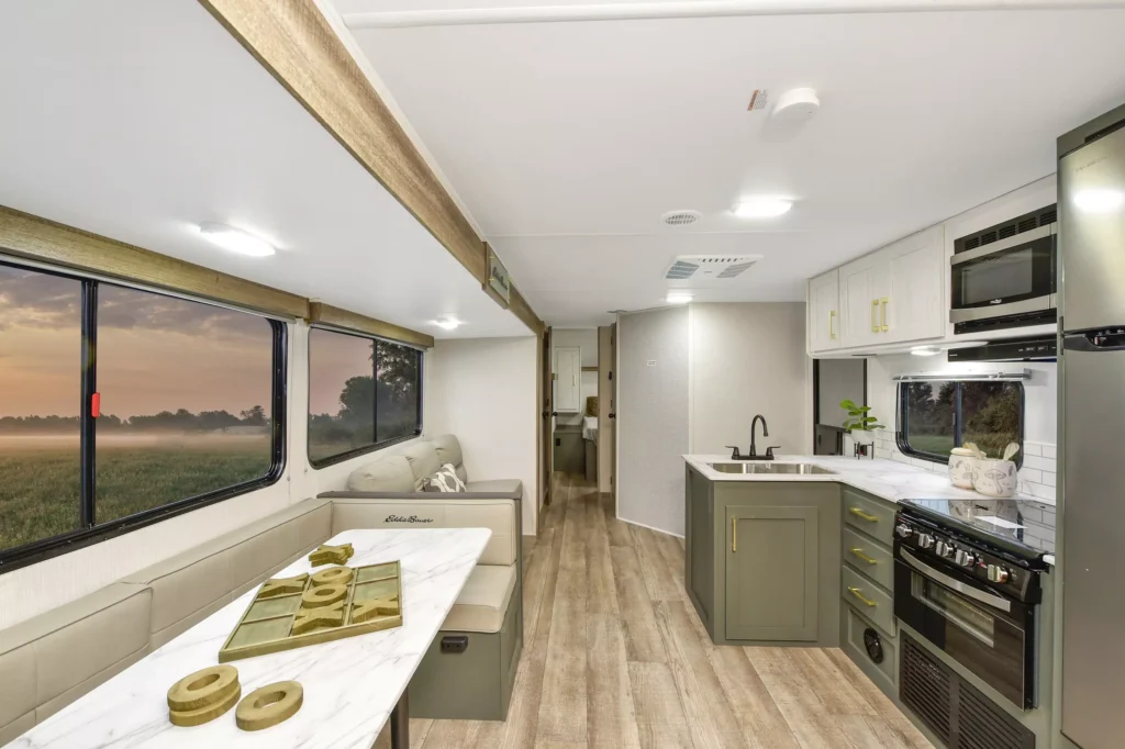 Modern RV interior with a kitchen, dining area, and sofa. Large window to the left shows a scenic sunrise over a field. Light wood floors, white cabinetry, and green accents create a clean, stylish space.