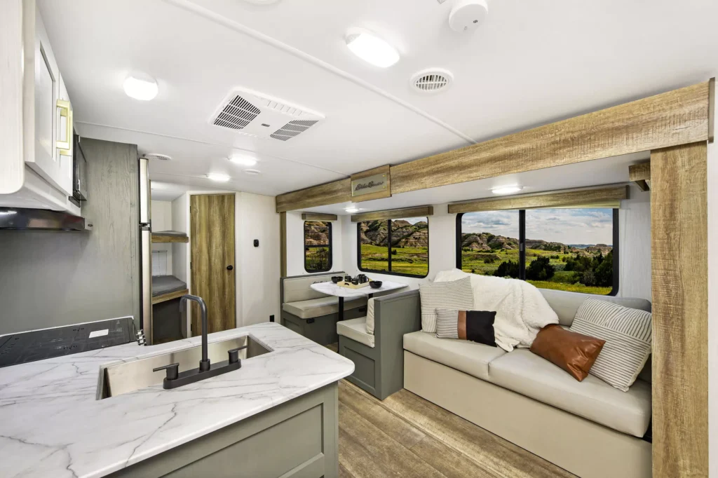 Modern RV interior with light wood floors, marble countertops, a kitchen sink, a dining table with bench seating, a sofa with pillows and blankets, large windows showing green landscape, and a doorway to the bedroom area.