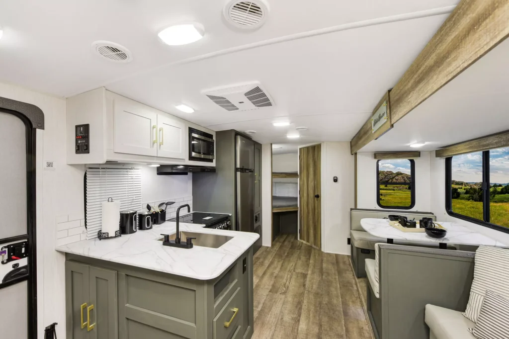 Modern RV interior with a marble-look countertop, kitchenette with appliances, wood-style flooring, and a dining area with large windows showing a scenic outdoor view. Bunk beds are visible in the background.