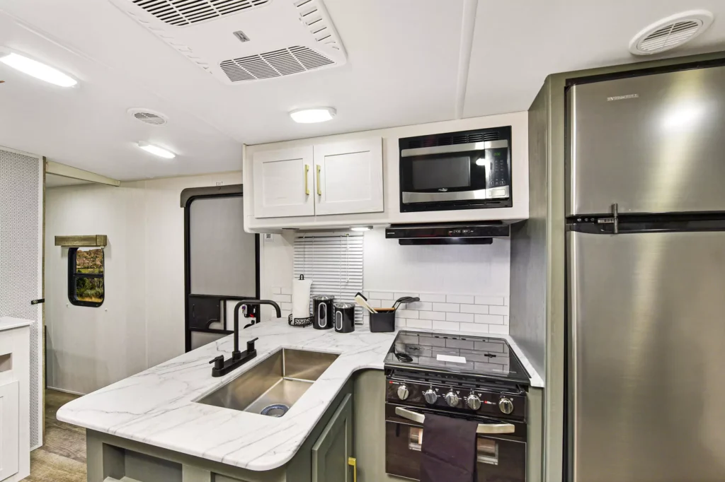 Modern RV kitchen with marble countertops, a stainless steel sink, black faucet, stove with oven, microwave, toaster, coffee maker, and a large stainless steel refrigerator. White cabinets and overhead lighting brighten the space.