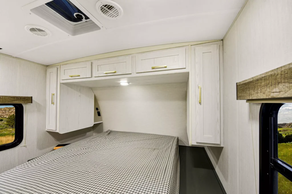 Interior of an RV bedroom with a checkered bedspread, white overhead cabinets, and two windows showing an outdoor landscape with grass and blue sky.