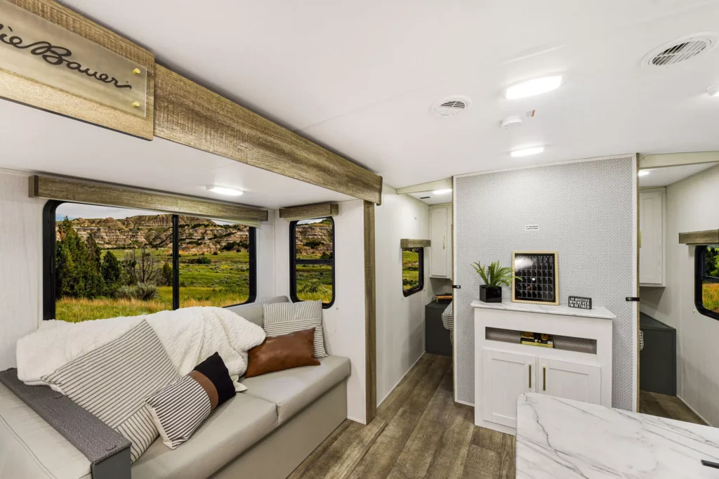 Modern RV interior with a light gray sofa, assorted cushions, and a throw blanket. Large windows reveal a scenic landscape. There’s a white fireplace below a wall shelf with plants and decor, and wood flooring throughout.