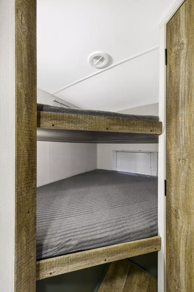 Two wooden bunk beds with gray mattresses in a small, light-colored room. The top and bottom bunks are stacked, and there is a window with closed blinds next to the beds. A wooden door is partially open on the right.