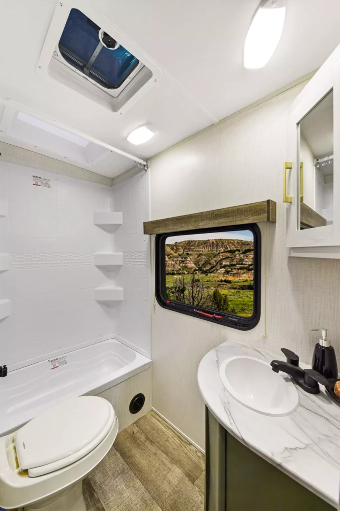 Modern RV bathroom with a toilet, bathtub, and marble sink. A large window shows a scenic outdoor view of rocky hills and blue sky. The interior is clean and well-lit with white walls and wood accents.