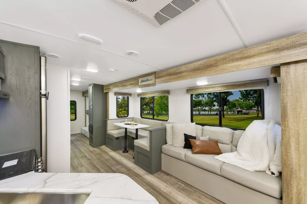Interior of a modern RV with a light-colored sofa, throw pillows, a blanket, a dining nook, large windows with views of trees and grass outside, and wood-style flooring and cabinetry.