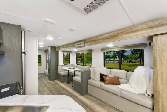 Interior of a modern RV with a light-colored sofa, throw pillows, a blanket, a dining nook, large windows with views of trees and grass outside, and wood-style flooring and cabinetry.