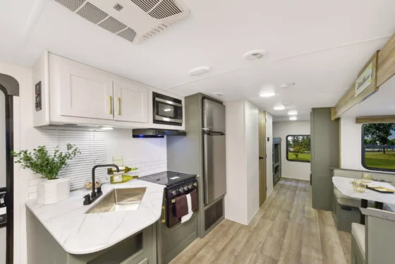 Interior of a modern RV kitchen with white cabinets, a stovetop oven, microwave, sink, and dining area. Large windows provide natural light, and a hallway leads to the sleeping area.