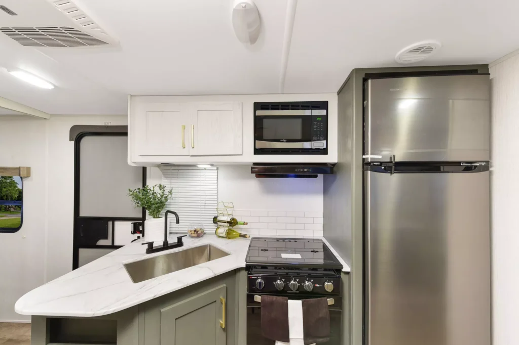 Modern RV kitchen with a marble-look countertop, sink, potted plant, stove, oven, microwave, and a stainless steel refrigerator. White upper cabinets and green lower cabinets add contrast.