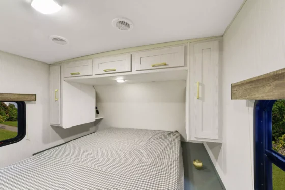 A small RV bedroom with a bed covered in a checkered blanket, overhead white cabinets, and a side window showing greenery outside. A round decorative item sits on a ledge beside the bed.