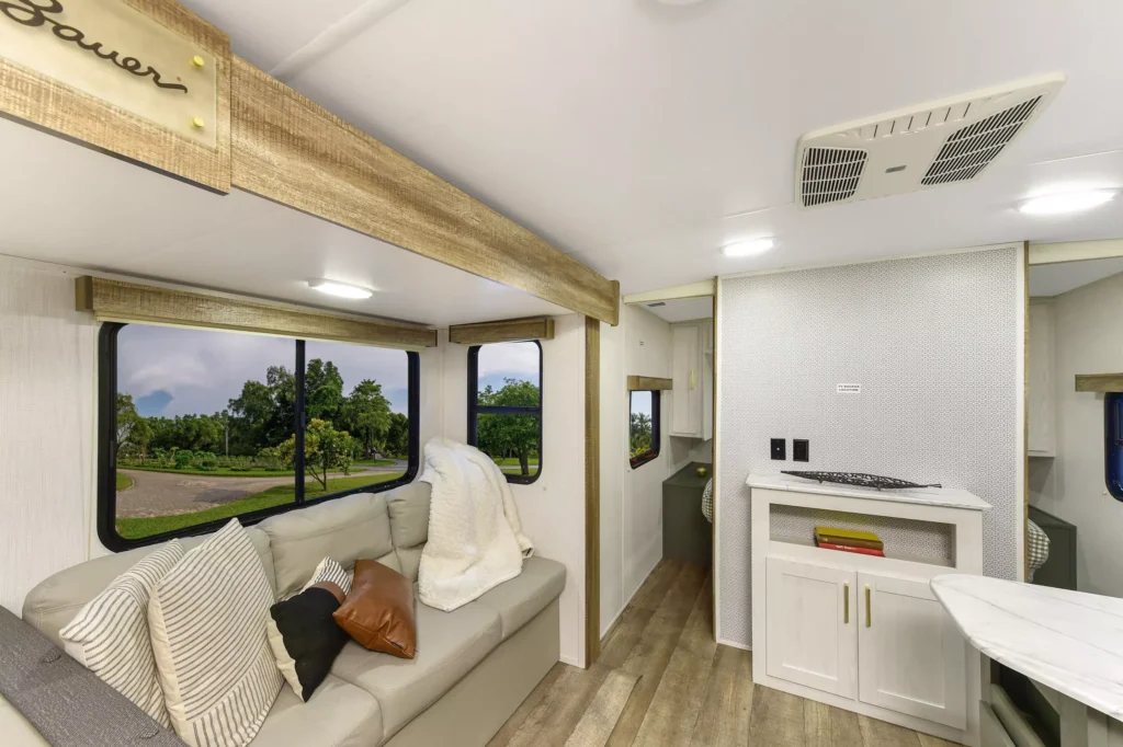 The image shows the interior of an RV with a beige sofa, assorted cushions, and a blanket. Large windows offer a view of greenery outside. Light wood flooring and cabinetry create a modern, cozy atmosphere.