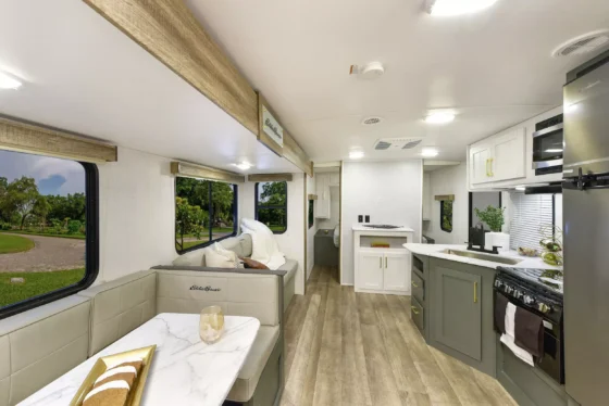 Modern RV interior featuring a dining area with a marble-style table, a sofa, kitchen with green and white cabinets, stove, microwave, and large windows showing greenery outside. Light wood flooring throughout.