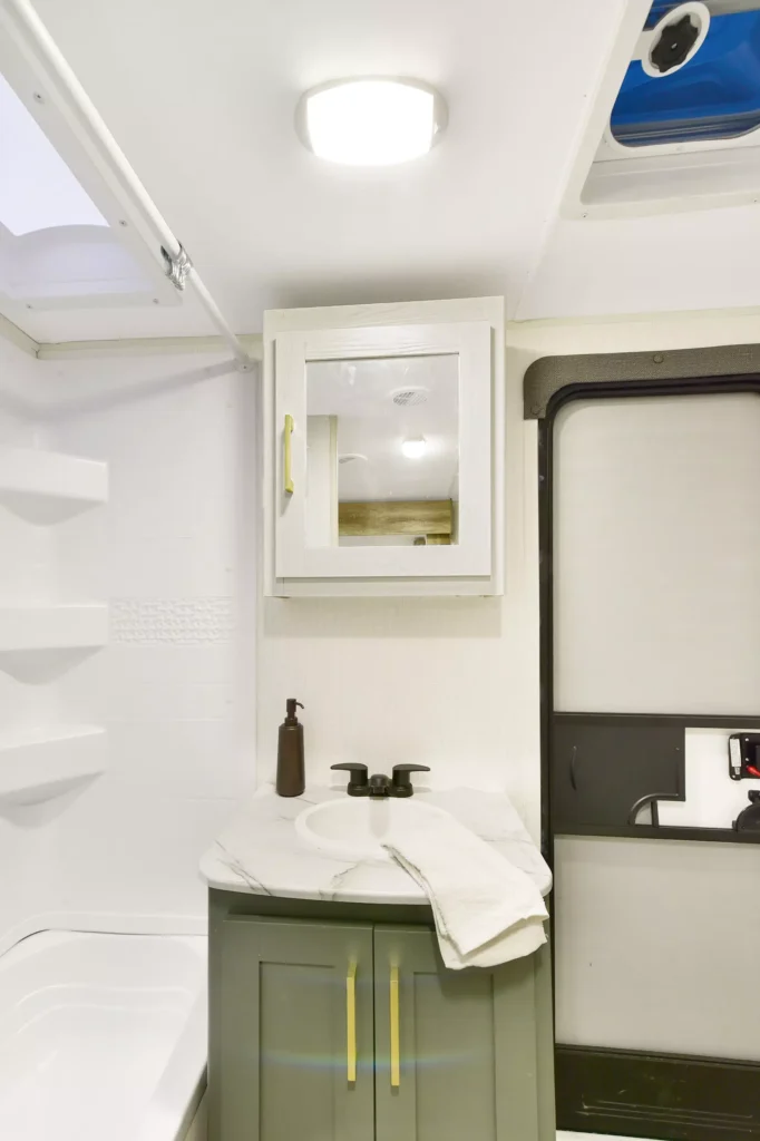A small bathroom with a gray vanity, white sink, countertop with towels and soap dispenser, mirrored medicine cabinet, bathtub with shelves, and an exterior door. Ceiling lights and a skylight provide illumination.