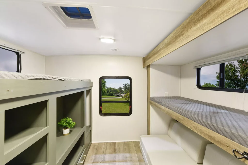 Interior of a camper with bunk beds, a small potted plant on a shelf, light wood and white walls, large window showing green grass and trees, and a skylight in the ceiling.