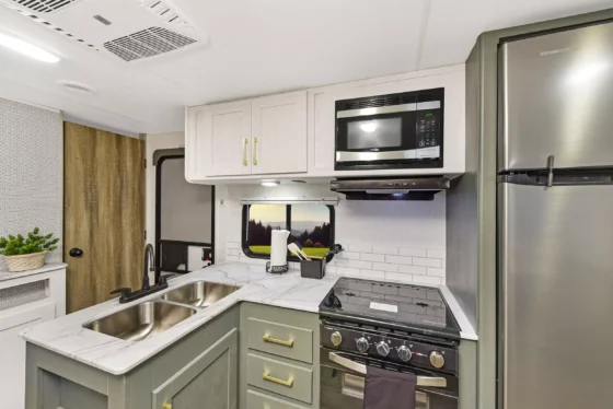 Modern RV kitchen with marble countertops, double sink, stainless steel fridge, oven with stove, microwave, light cabinets, backsplash tiles, potted plant, and a window above the sink.