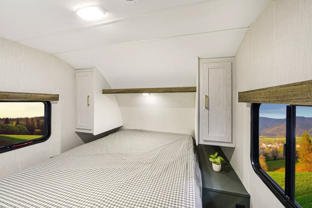 A cozy camper bedroom with a checkered bedspread, two windows showing scenic countryside views, overhead cabinets, and a small plant on a bedside table.