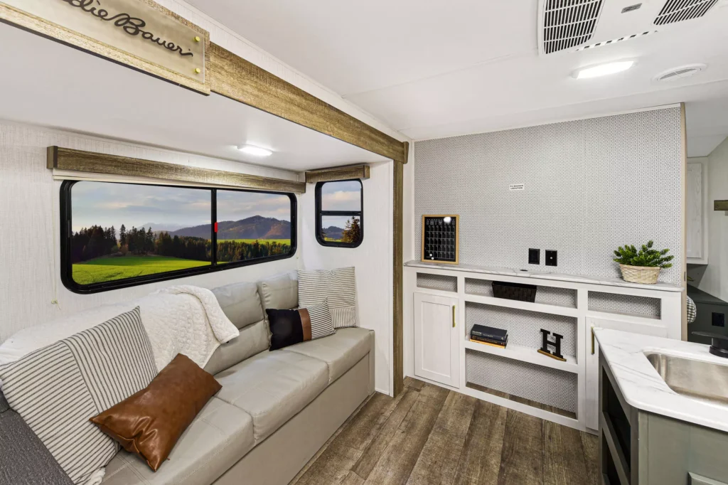 Interior of an RV with a beige sofa, pillows, a large window showing mountains and trees, wooden flooring, shelves with decor items, a small plant, and modern light fixtures.
