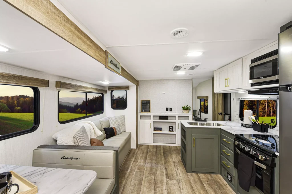 Modern RV interior with light wood floors, a kitchen with green cabinets, stainless steel appliances, a dining table, and a cozy seating area. Large windows show a scenic view of green fields and trees outside.