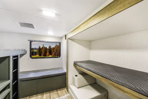 A clean, modern RV interior features two stacked bunk beds with gray bedding, light wood and neutral walls, and a window showing a scenic view of trees and mountains outside.