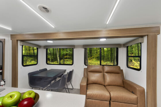 A modern RV interior with tan leather seating, a black dining table with black chairs, large windows showing a green forest, and a counter with a bowl of apples in the foreground.