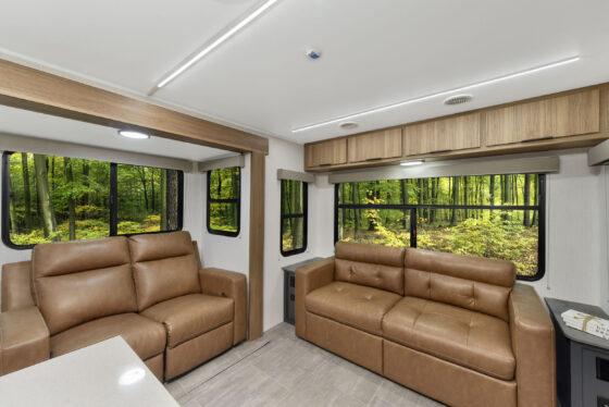 Interior of an RV with two brown leather sofas, large windows showing a green forest outside, light-colored wood cabinets, and neutral-colored flooring and walls.