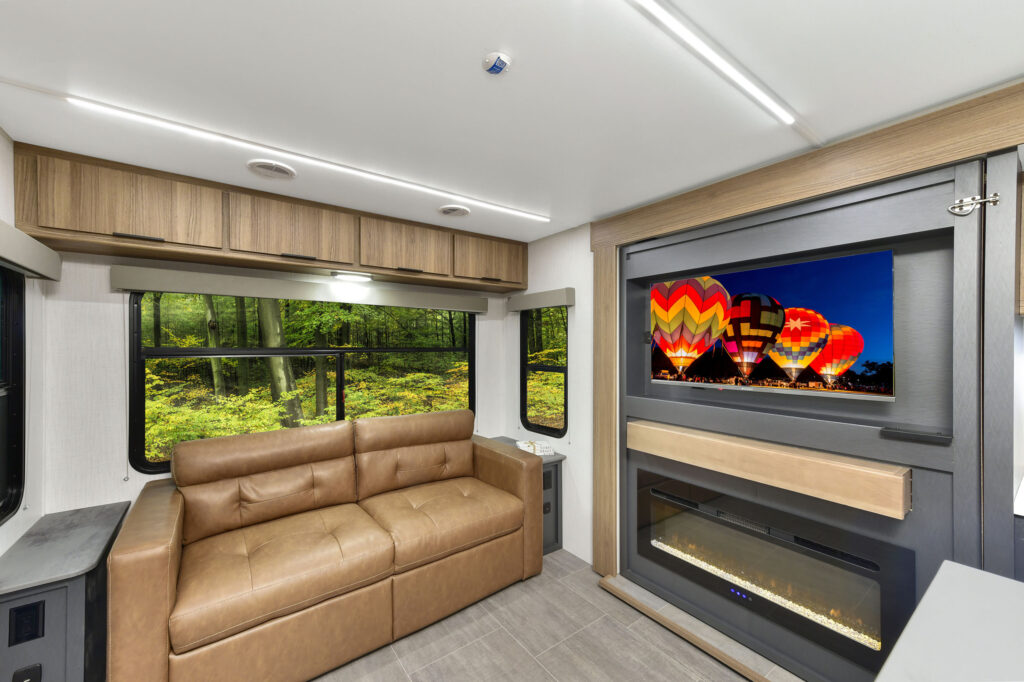 Modern RV interior featuring a tan leather sofa, large window with a forest view, built-in electric fireplace, and a wall-mounted TV displaying colorful hot air balloons.
