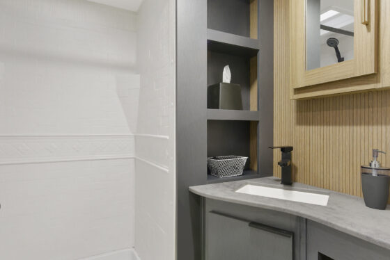 Modern bathroom with a white shower on the left, dark gray shelving and sink cabinet in the center, and a wooden medicine cabinet with a mirror on the right. The color scheme is neutral and minimalist.