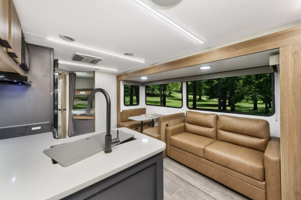 Modern RV interior with a tan leather sofa, built-in dining area, large windows showing green trees, and a sleek kitchen featuring a sink and white countertops. Bright lighting and wood accents create an inviting atmosphere.