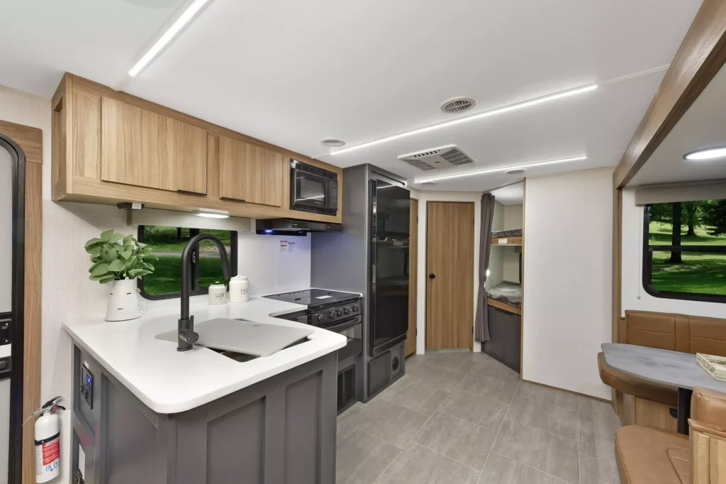 Modern RV interior featuring a kitchen with a sink, stove, microwave, wooden cabinets, a small dining area, and a view of a bunk bed and bathroom door; large window shows green park outside.