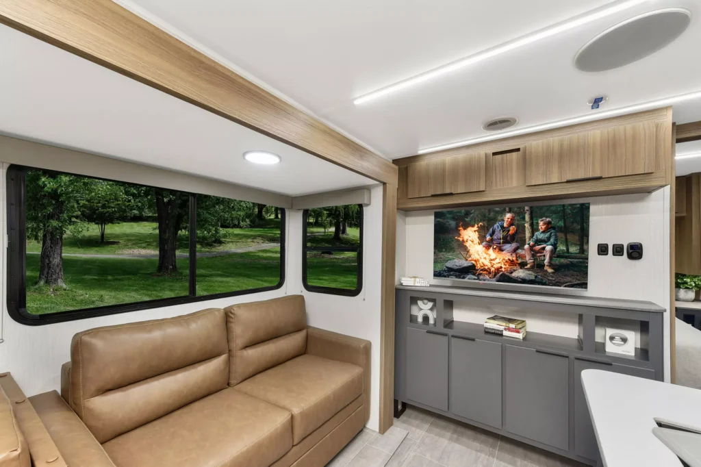 The interior of a modern RV features a brown leather sofa, large windows with a view of green trees, and a wall-mounted TV displaying a group gathered around a campfire. Shelves and cabinets provide storage.