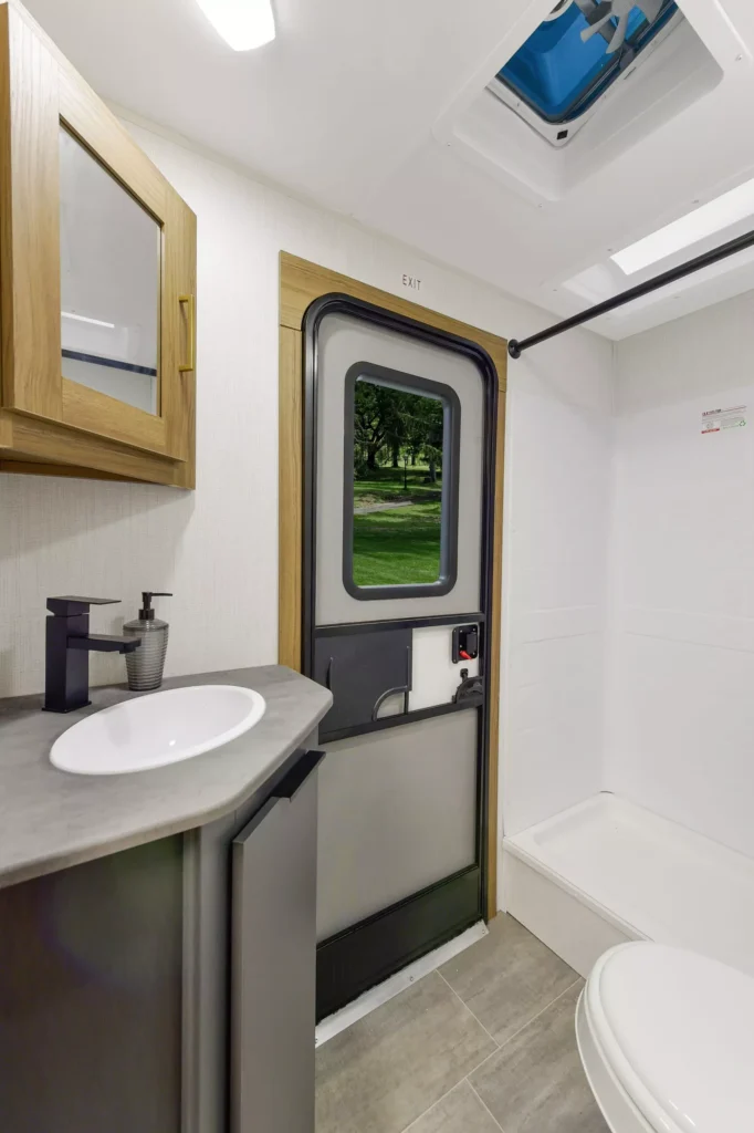 A compact RV bathroom with a sink and mirrored cabinet on the left, a toilet and shower on the right, and a door with a window showing a green park and trees outside.