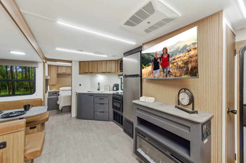 Modern RV interior with kitchen, dining area, and bed in background. A wall-mounted TV displays two people hiking. The space features light wood accents, gray cabinetry, and an electric fireplace with a clock on top.
