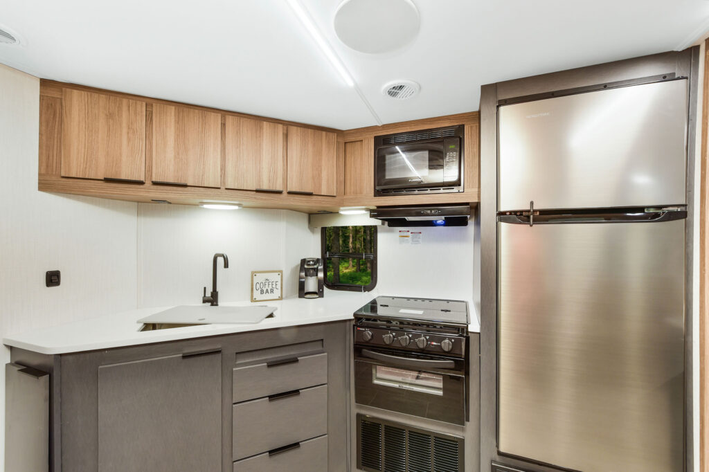 A modern RV kitchen with wood cabinets, a stainless steel fridge, oven, microwave, and a sink. A coffee maker and a sign reading Coffee Bar sit on the white countertop.