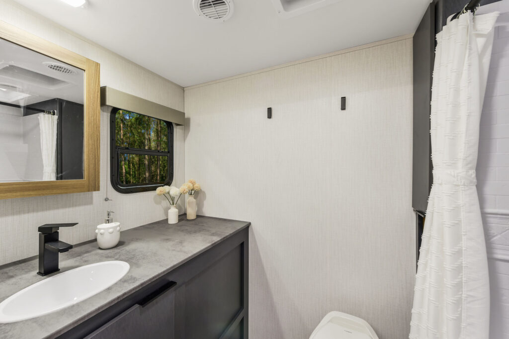 Modern RV bathroom with a gray countertop, black faucet, white sink, and decorative flowers beside a mirror. There are hooks on the beige wall and a window showing trees outside. A shower with a white curtain is on the right.
