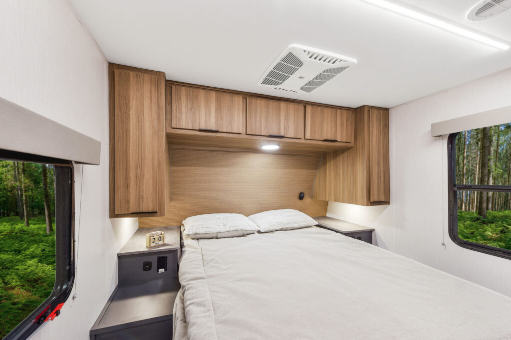 Interior of a modern RV bedroom with a neatly made bed, light wood cabinets above, nightstands on each side, windows showing a forest view, and a small clock on the left nightstand.