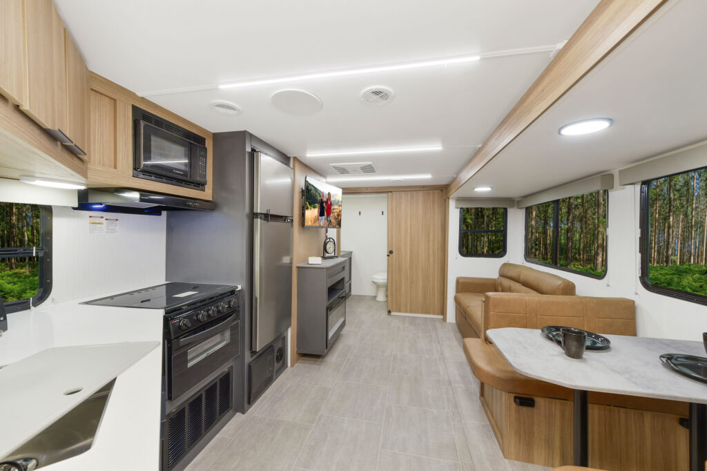 Modern RV interior featuring a kitchen with stove and microwave, a dining booth, a sofa, a mounted TV, and large windows showing a wooded outdoor scene. A bathroom is visible at the far end.