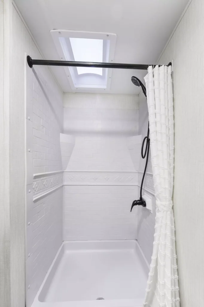 A small white shower with a textured white curtain, a black showerhead mounted on the wall, a white tiled interior, and a skylight overhead providing natural light.
